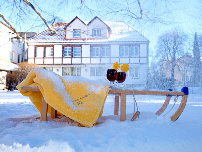 Ein mit Schnee bedeckter Holzschlitten, auf dem zwei Getränke stehen, vor dem charmant-rustikalen Gebäude des Braunschweiger Hofs in Niedersachsen.